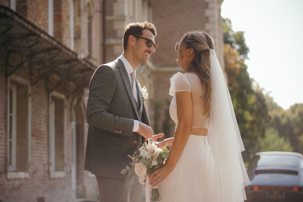 Plan rapproché du couple Alizée et son époux. La mariée porte une robe de mariée deux-pièces sur mesure de Maïté Bailleul, devant le Château de Flixecourt.