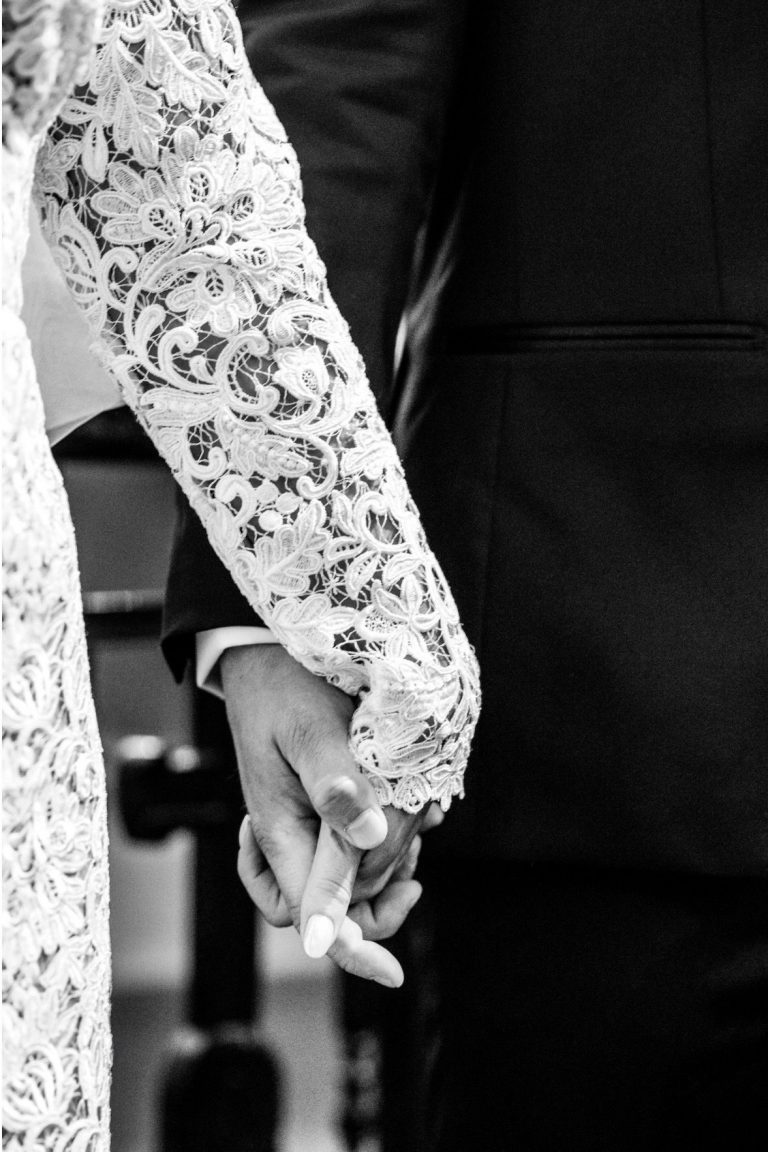 Gros plan en noir et blanc des mains des mariés entrelacées. On voit le détail de la manche en dentelle de la mariée et la main du marié tenant la sienne