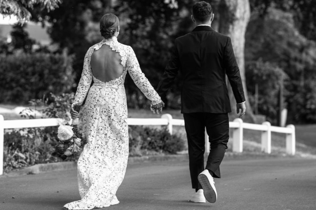 Vue de dos des mariés Marie et Dan marchant main dans la main dans un parc. La mariée porte une robe en dentelle à manches longues et un grand dos nu.