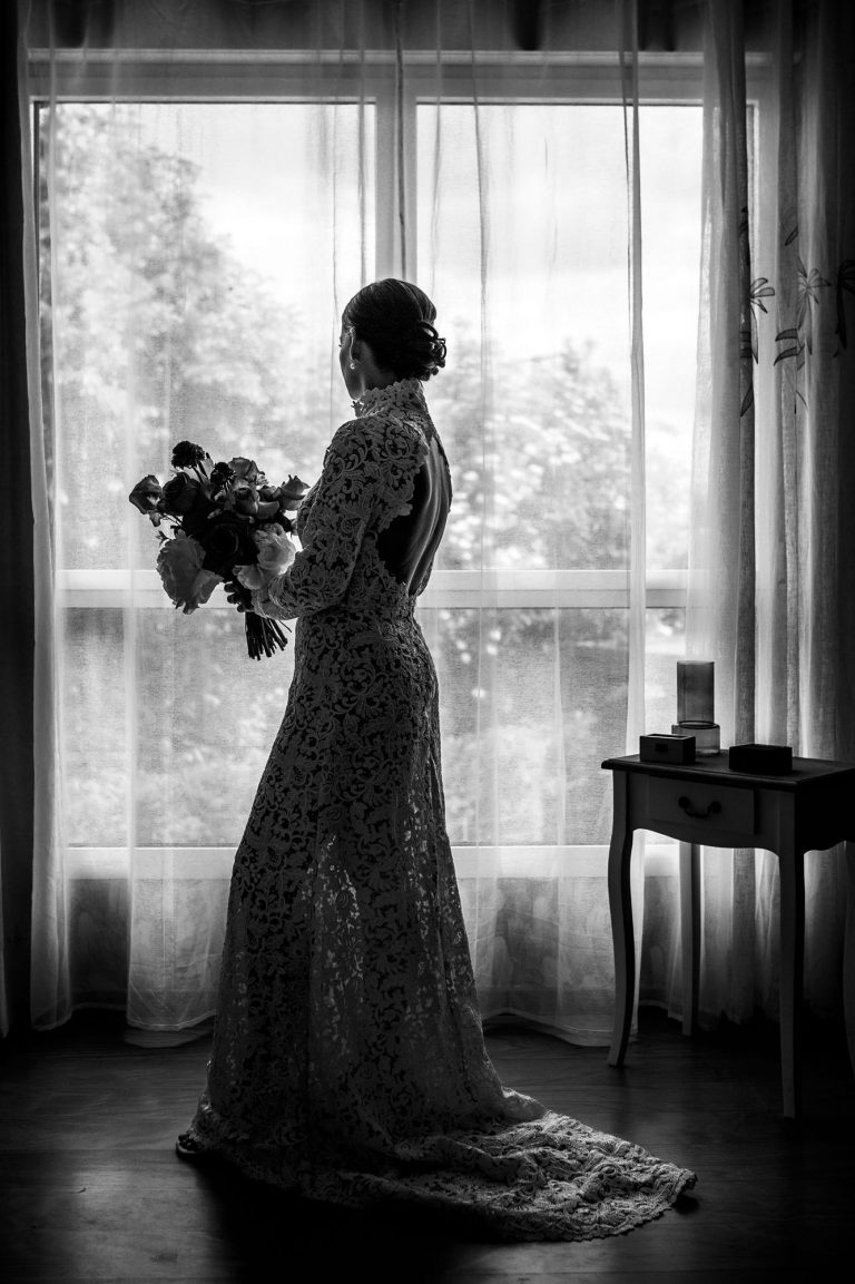 Silhouette de la mariée Marie vue de profil, tenant son bouquet devant une fenêtre. Sa robe en dentelle met en valeur le dos nu et la traîne.