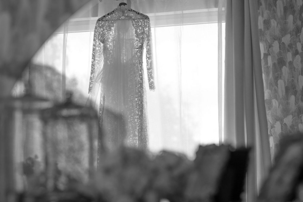 Gros plan de la robe de mariée en dentelle à manches longues, suspendue devant une fenêtre lumineuse avant la cérémonie, mettant en valeur le tissu fin.